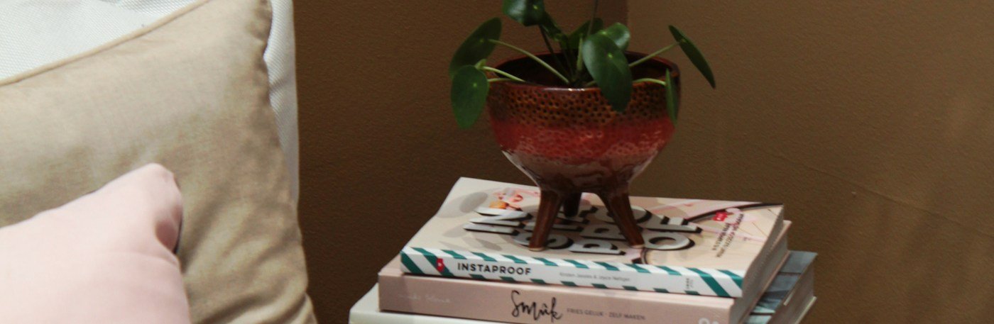 Un coin de lecture, trois livres empilés sur un tabouret de bois, une plante en pot, des coussins et une couverture